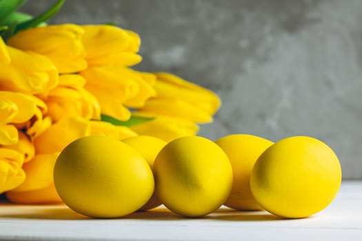 Bouquet of bright yellow tulips and Easter eggs lying on white wooden surface on gray background, copy space