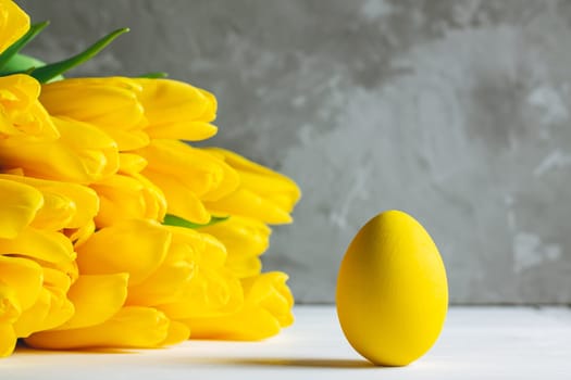 Bouquet of bright yellow tulips and Easter egg, standing on white wooden surface on gray background, copy space