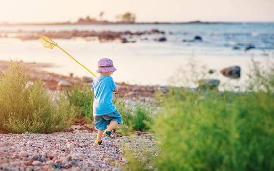 Child going to the water with net in his hand along pebble seaside. Concept of the family vacation and tourism.