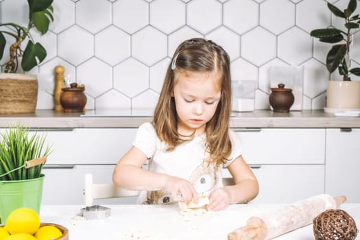 Portrait of little serious girl, helping, making shape dough easter cookies. Sculpt dough flour bakery cup biscuit. Preparing rolling pin. Wood plate of yellow eggs, plant. Kitchen utensil indoor.