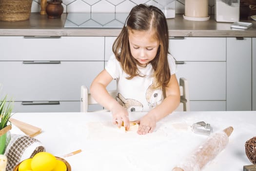 Portrait of little serious girl, helping, making shape dough easter cookies. Sculpt dough flour bakery cup biscuit. Preparing rolling pin. Wood plate of yellow eggs, plant. Kitchen utensil indoor.