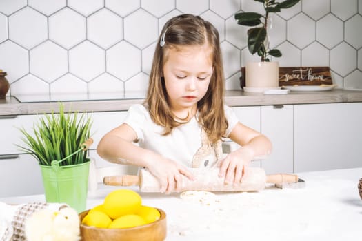Portrait of little serious girl, helping, learning rolling dough, making shape easter cookies. Sculpt flour biscuit. Preparing holding rolling pin. Yellow egg, plant. Kitchen utensil indoor. Light day