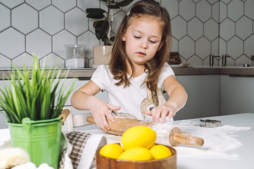 Portrait of little serious girl, helping, learning rolling dough, making shape easter cookies. Sculpt flour biscuit. Preparing holding rolling pin. Yellow egg, plant. Kitchen utensil indoor. Light day