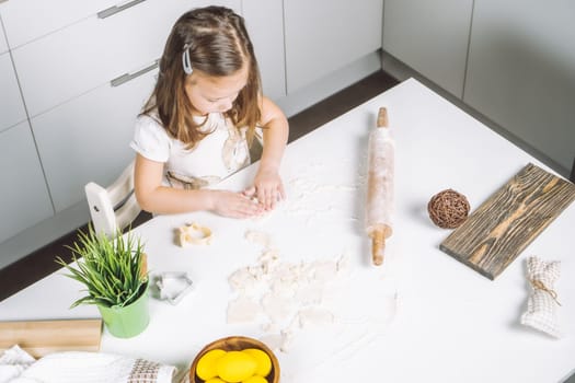 Portrait of little serious girl, helping, making shape dough easter cookies. Sculpt dough flour bakery cup biscuit. Preparing rolling pin. Wood plate of yellow eggs, plant. Kitchen utensil indoor.
