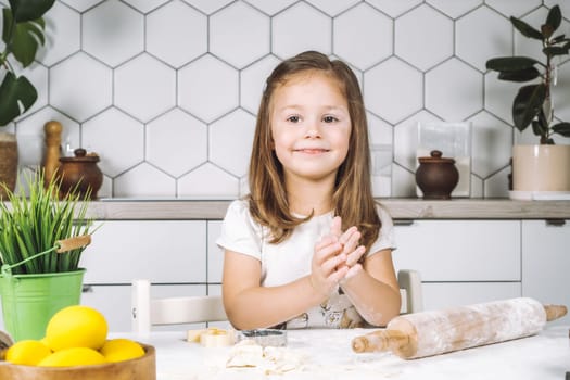 Portrait of little smiling girl sitting on chair, kneading, making different shape cookies. Preparing dough, rolling pin with flour. Sculpting shape biscuit. Plate yellow easter eggs, plant pot