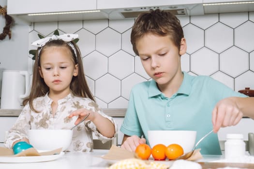 Portrait of concentrated two little children, boy, girl, multi coloring Easter hens eggs. Soaking up, absorption color animal eggs spoon, bowls. Paper on table in kitchen. Traditional holiday event.
