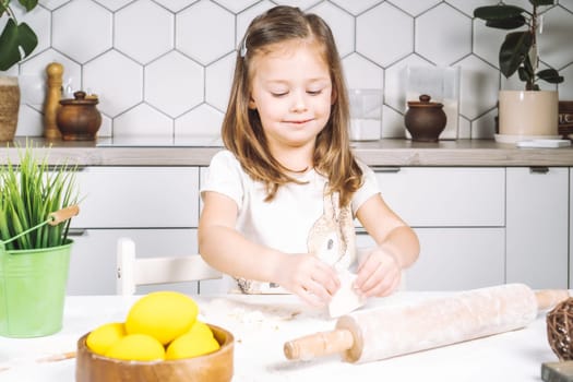 Portrait of little studiously girl sitting on chair, assistance, making different shape cookies. Preparing dough, rolling pin with flour. Sculpting shape biscuit. Plate yellow easter eggs, plant pot