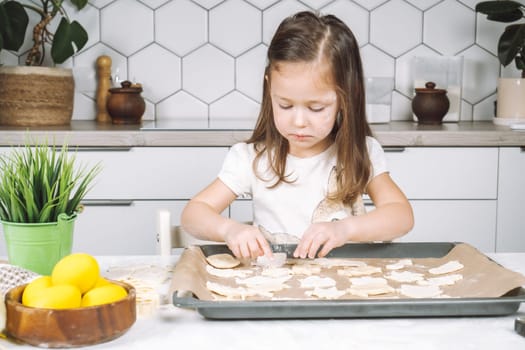 Portrait of little studiously girl chef, sitting on chair, making different shape dough cookies on dripping pan. Wooden plate of yellow easter eggs and plant pot near child. Kitchen utensil indoor.