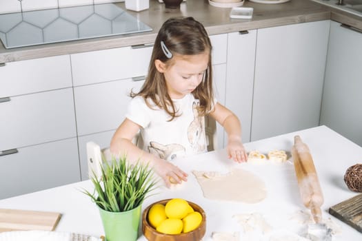 Portrait of concentrated little girl chef, sculpting dough shape cookies in baking cup. Sitting on chair kitchen, Wooden plate of yellow easter eggs and plant pot near child. Kitchen utensil indoor.