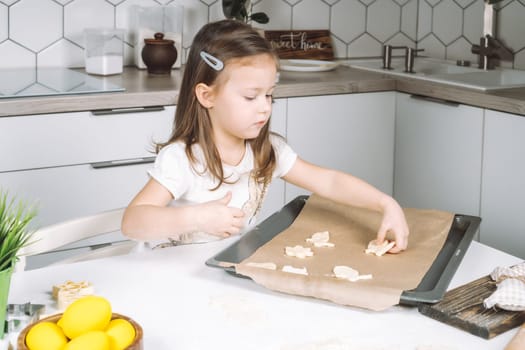 Portrait of little studiously girl chef, sitting on chair, making different shape dough cookies on dripping pan. Wooden plate of yellow easter eggs and plant pot near child. Kitchen utensil indoor.