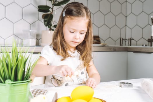 Portrait of little serious girl, helping, making shape dough easter cookies. Sculpt dough flour bakery cup biscuit. Preparing rolling pin. Wood plate of yellow eggs, plant. Kitchen utensil indoor.