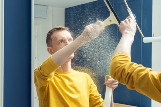 Middle aged man spray cleanser on room mirror and wipe splashes with plastic brush. Rub from dust and polish mirror with household tool. Clear house, housework, wet home cleaning concept.