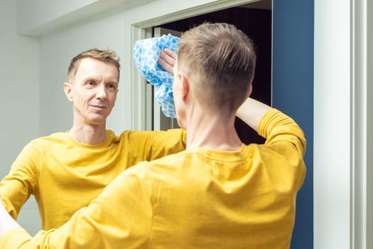 Smiling mature man wiping room mirror with dry rag, back view. Rub from dust and polish mirror with household cloth. Domestic life, tidy housekeeper. Housework, home cleaning concept.