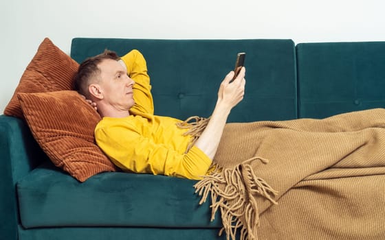 Adult man lie on sofa and watch video on smartphone in living room, side view. Person reclining on pillow under blanket and lounging. Look at screen of phone, read and chat. Home relaxation, leisure.