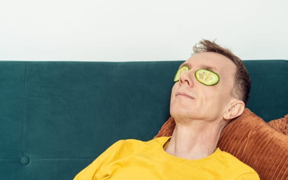 Happy man lying on sofa with eye patches from fresh cucumber slices in living room. Adult person reclining on pillow with mask for dark circles under eyes. Home relaxation, beauty treatment.