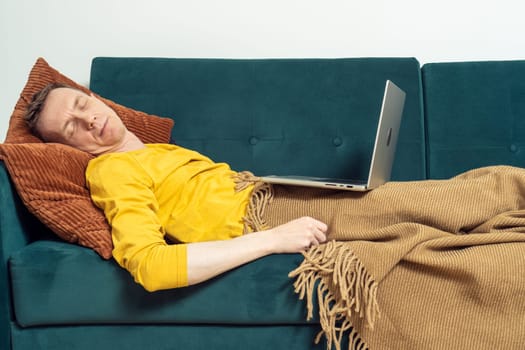 Tired man sleeping on sofa with open laptop in living room. Overworking person reclining on pillow under blanket. Home relaxation, rest break. Business people, remote work, freelancing.