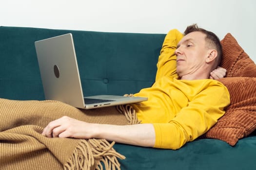 Tired man lie on sofa and watch movie on laptop in living room. Adult person reclining on pillow under blanket. Look at monitor of notebook and read. Home relaxation, leisure, lazy activity.