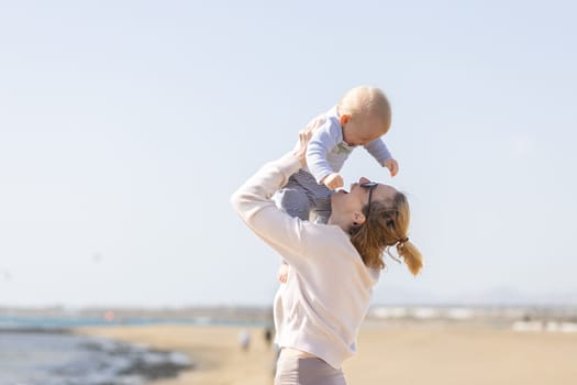 Mother enjoying summer vacations holding, playing and lifting his infant baby boy son high in the air on sandy beach on Lanzarote island, Spain. Family travel and vacations concept