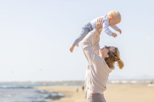 Mother enjoying summer vacations holding, playing and lifting his infant baby boy son high in the air on sandy beach on Lanzarote island, Spain. Family travel and vacations concept