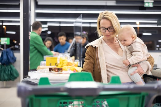 Mother shopping with her infant baby boy, holding the child while stacking products at the cash register in supermarket grocery store