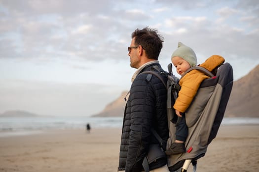 Father rising hands to the sky while enjoying pure nature carrying his infant baby boy son in backpack on windy sandy beach of Famara, Lanzarote island, Spain at sunset. Family travel concept