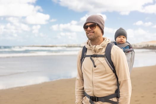 Young father carrying hisinf ant baby boy son in backpack on windy sandy Famara beach, Lanzarote island, Spain. Family travel and winter vacation concept