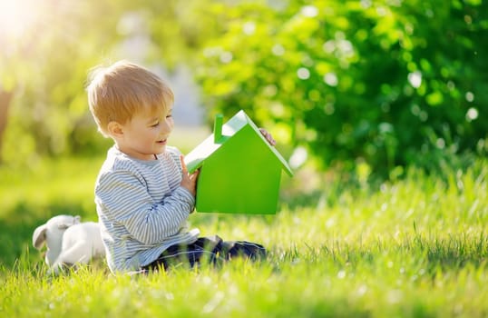 Smiling child sitting on the spring meadow with model of the green house in his hands. Concept of the real estate, rental and buying of a new apartment.