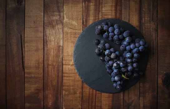 Blue Isabella grapes bunch and round stone slate plate on on wooden background. Copy space for text. Flat lay.
