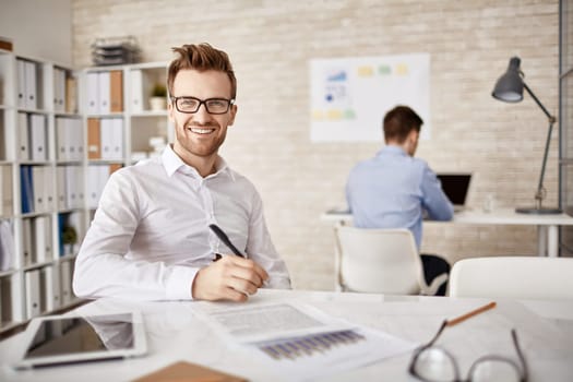 Successful young businessman sitting by workplace and looking at camera