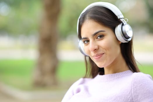 Happy woman with headphones looks at you in a park