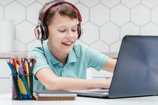 Portrait of young preteen exciting boy wearing T-shirt, headphones, sitting at desk in front of laptop, typing, chatting, playing games, messaging, doing homework. Gaming, e-learning, social media.