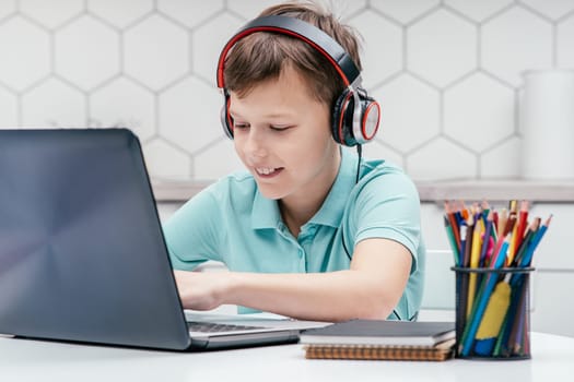 Portrait of young preteen concentrated boy wearing T-shirt, headphones, sitting at desk in front of laptop, typing chatting, playing games, messaging, communicating. Gaming, e-learning, social media.