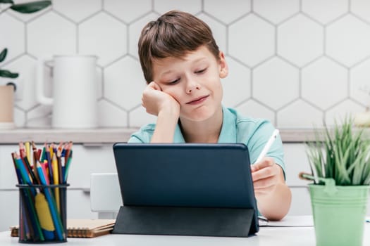 Portrait of young preteen pretty boy wearing blue T-shirt, sitting at desk in front of digital tablet, writing drawing on screen with white pen stylus. Remote education, online school, e-learning.