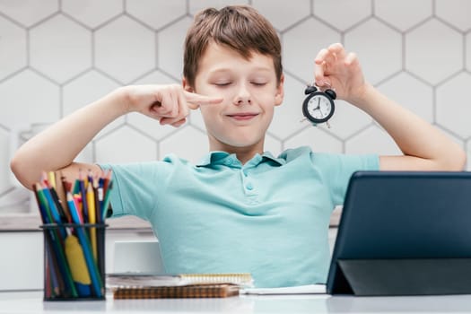 Portrait of young preteen happy boy with closed eyes wearing blue T-shirt, sitting at desk with pencils, digital tablet, pointing index finger at small clock. Education, school, e-learning, deadline.