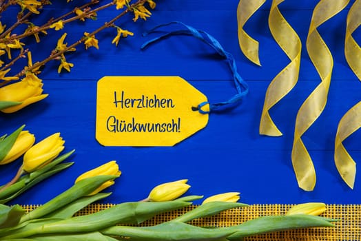 Label With German Text Herzlichen Glueckwunsch Means Congratulations. Yellow Spring Flowers Like Tulip And Branches. Festive Decoration With Ribbon. Blue Wooden Background
