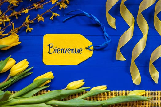 Label With French Text Bienvenue Means Welcome. Yellow Spring Flowers Like Tulip And Branches. Festive Decoration With Ribbon. Blue Wooden Background