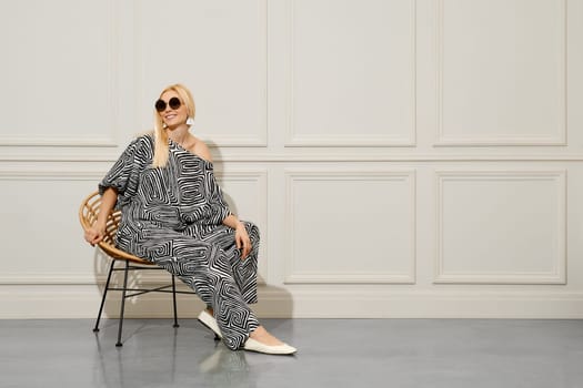 Happy blonde fashionable woman in blouse and loose trousers with twisted strips sits in armchair in the room