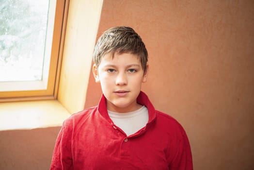 A white boy of school age stands near the window, dressed in a red shirt.