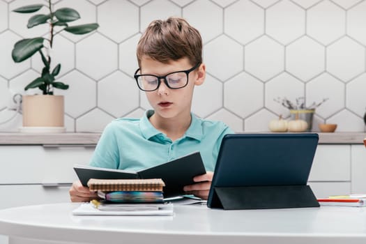 Portrait of young preteen focused boy wearing blue T-shirt, glasses, sitting at desk near notepads, looking at open black book near tablet, having online lesson, reading. E-learning, technology.