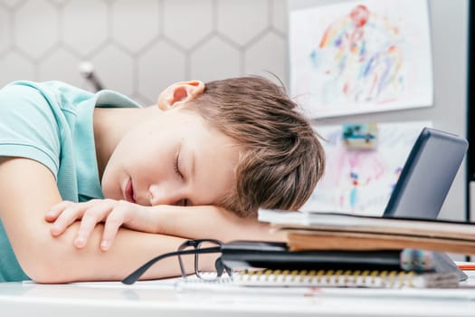 Side view of young preteen exhausted boy pupil wearing blue T-shirt, lying sleeping on folded hands on desk near notebooks, glasses, tablet at home. Distant learning, online education, technology.
