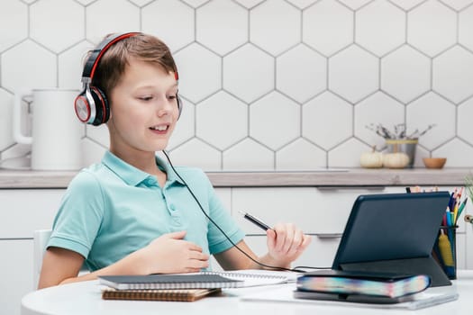 Portrait of young preteen busy boy wearing T-shirt, headphones, sitting at desk near notebooks, looking at screen of tablet, having online lesson, holding pen. Remote education, distance learning.
