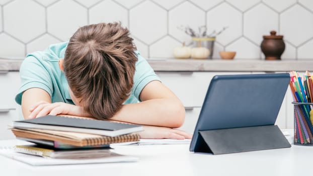 Portrait of young preteen tired boy pupil wearing blue T-shirt lying sleeping on folded hands on desk near notebooks, glasses, color pencils, tablet at home. Distant learning, education, technology.