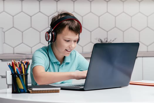 Portrait of young preteen pleased boy wearing T-shirt, headphones, sitting at desk near pencils, using laptop, playing games, messaging, listening to music. Gaming, distance learning, social media.