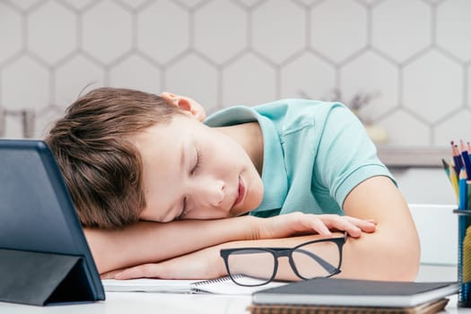 Portrait of young preteen exhausted boy wearing blue T-shirt, lying sleeping on hands on desk near notebooks, glasses, color pencils, tablet at home. Distant learning, homeschooling, technology.