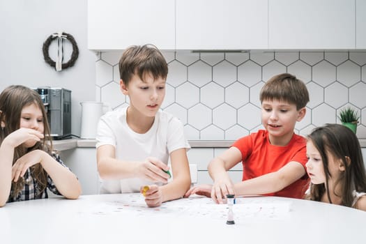 Happy little school friends play board game on paper map with dice and chips at kitchen table. Children throw dice and move figure. Home leisure, playtime. Entertaining interesting table game.
