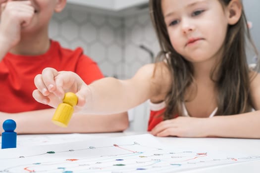 Little school friends play board game on paper map with dice and chips at kitchen table. Pretty girl hold in hand yellow figure closeup. Home leisure, playtime. Entertaining interesting table game.