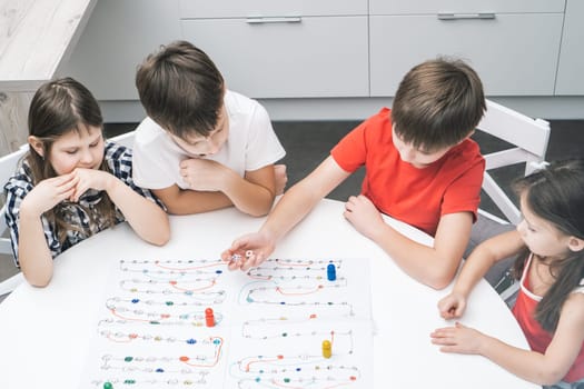 Little school friends play board game with dice and chips on kitchen table, from above view. Kids look at boy throw dice and move figure. Home leisure, playtime. Entertaining interesting table game.