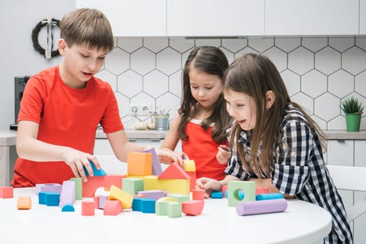 Funny school friends play toy constructor on kitchen table. Girls and boy collect colorful details of lego. Little kids build toy city. Home leisure, playtime. Educational interesting board game.