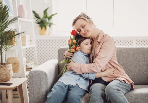 Cute boy sitting on the sofa with mom and giving a bouquet of tulips to her. Concept of the Mother's day and family relation.
