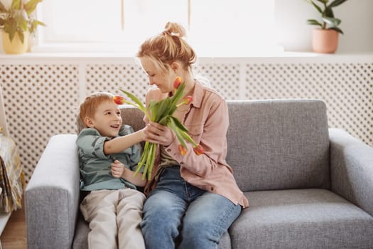 Cute boy sitting on the sofa with mom and giving a bouquet of tulips to her. Concept of the Mother's day and family relation.